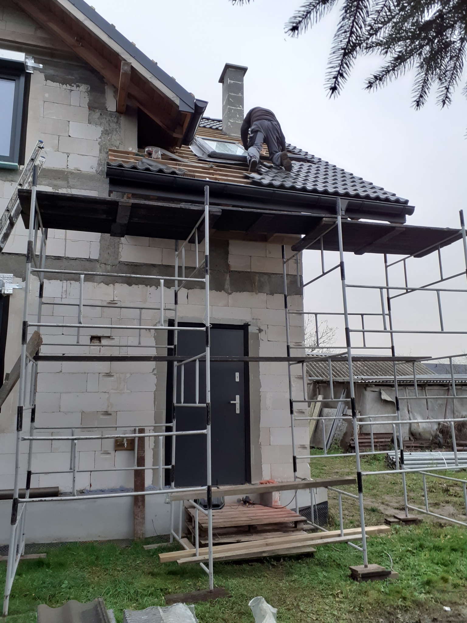 Prace dekarskie na dachu domu w trakcie budowy, widoczne rusztowanie, komin i okno dachowe.