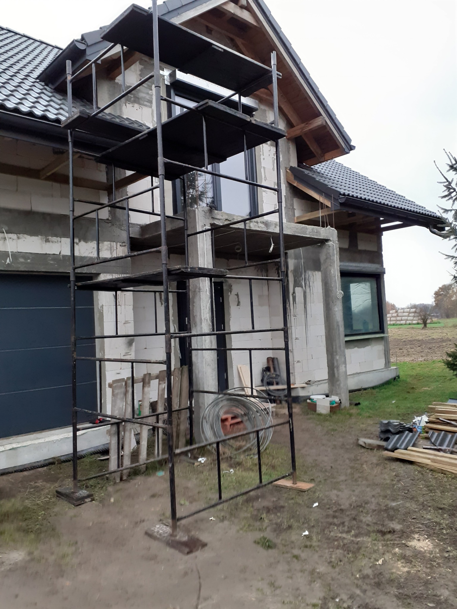 Dom w trakcie budowy z częściowo ukończonym dachem z czarnej dachówki, widoczne rusztowanie i materiały budowlane na zewnątrz.
