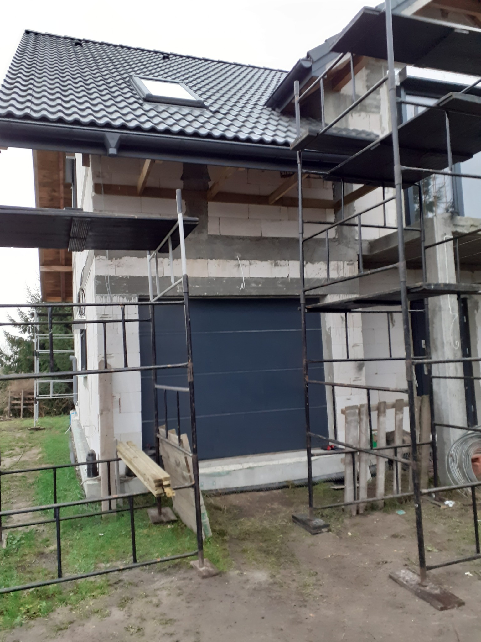 Dom w trakcie budowy z nowym dachem z dachówki ceramicznej, widoczne rusztowania i częściowo otynkowane ściany.