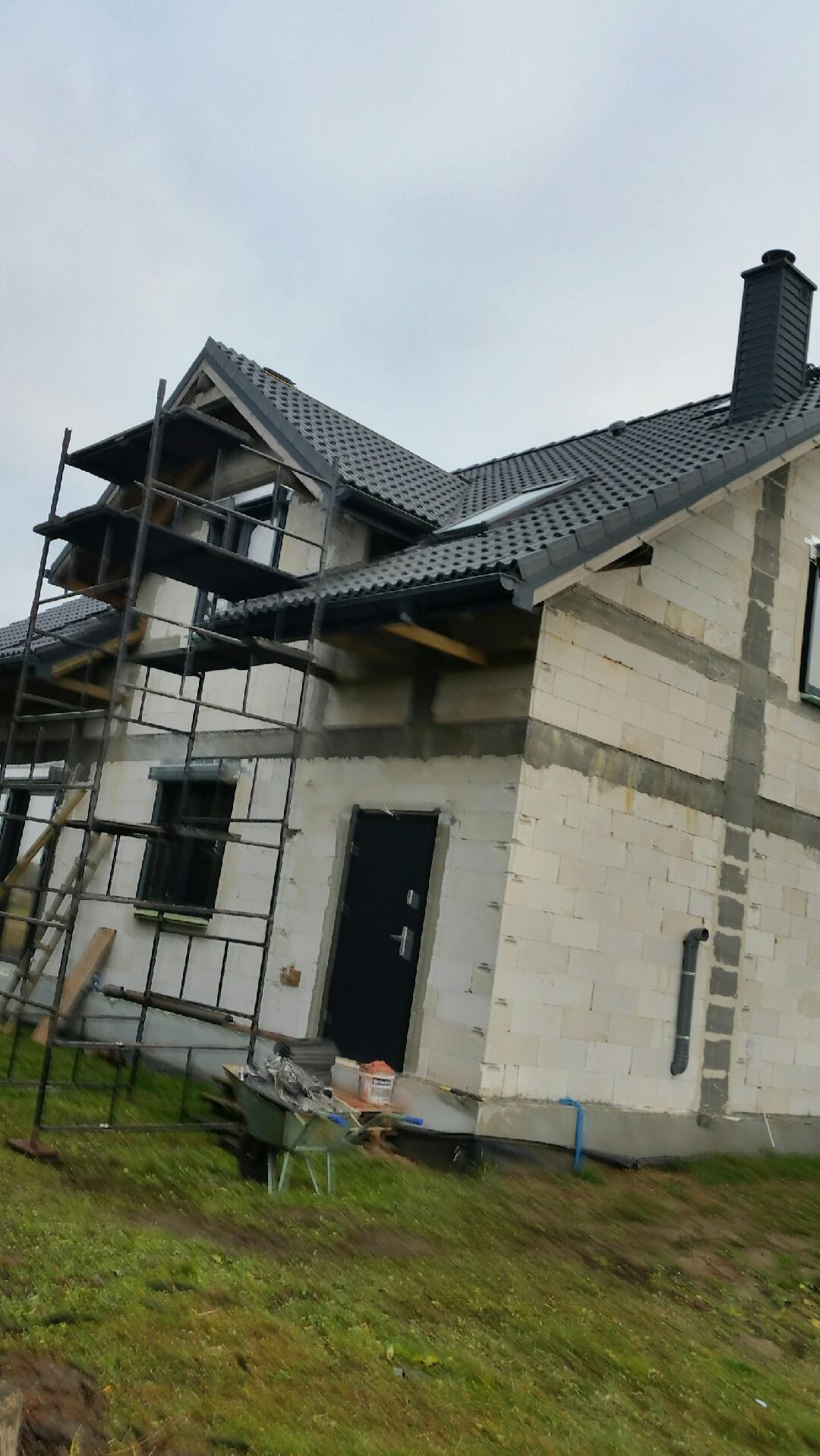 Surowy stan domu jednorodzinnego w trakcie budowy: szare dachówki na dachu, rusztowania przy ścianach z białego bloczka, czarne okna i drzwi.