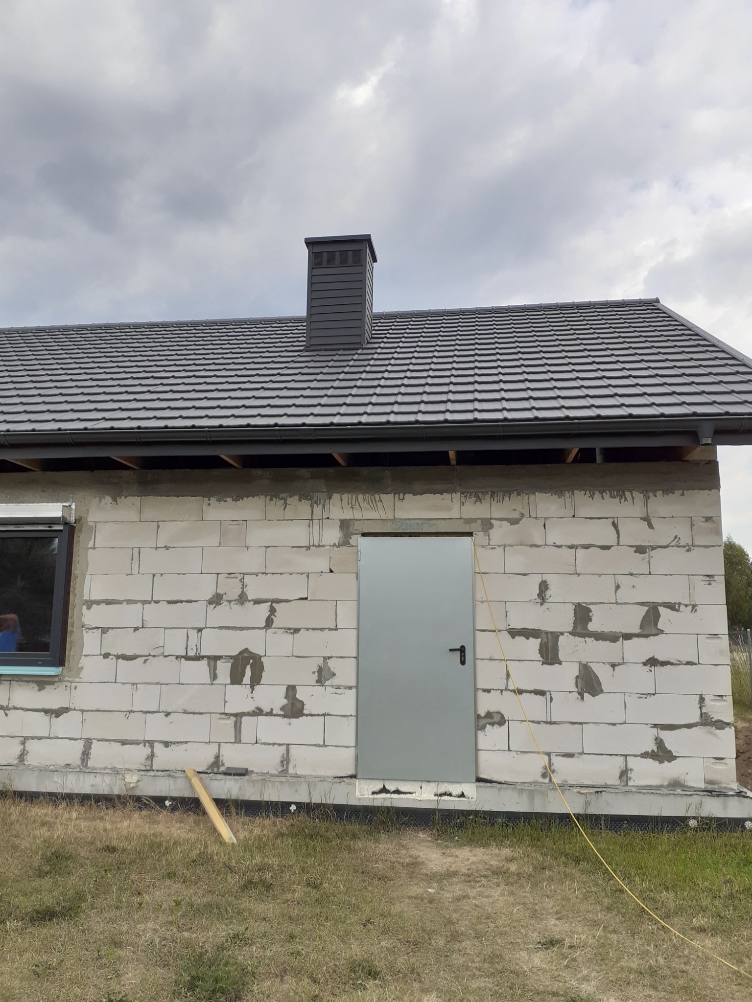 Surowy budynek w trakcie budowy z szarym dachem z dachówki ceramicznej, kominem i metalowymi drzwiami, widoczne bloczki betonowe i niezabezpieczone drewniane elementy pod dachem.