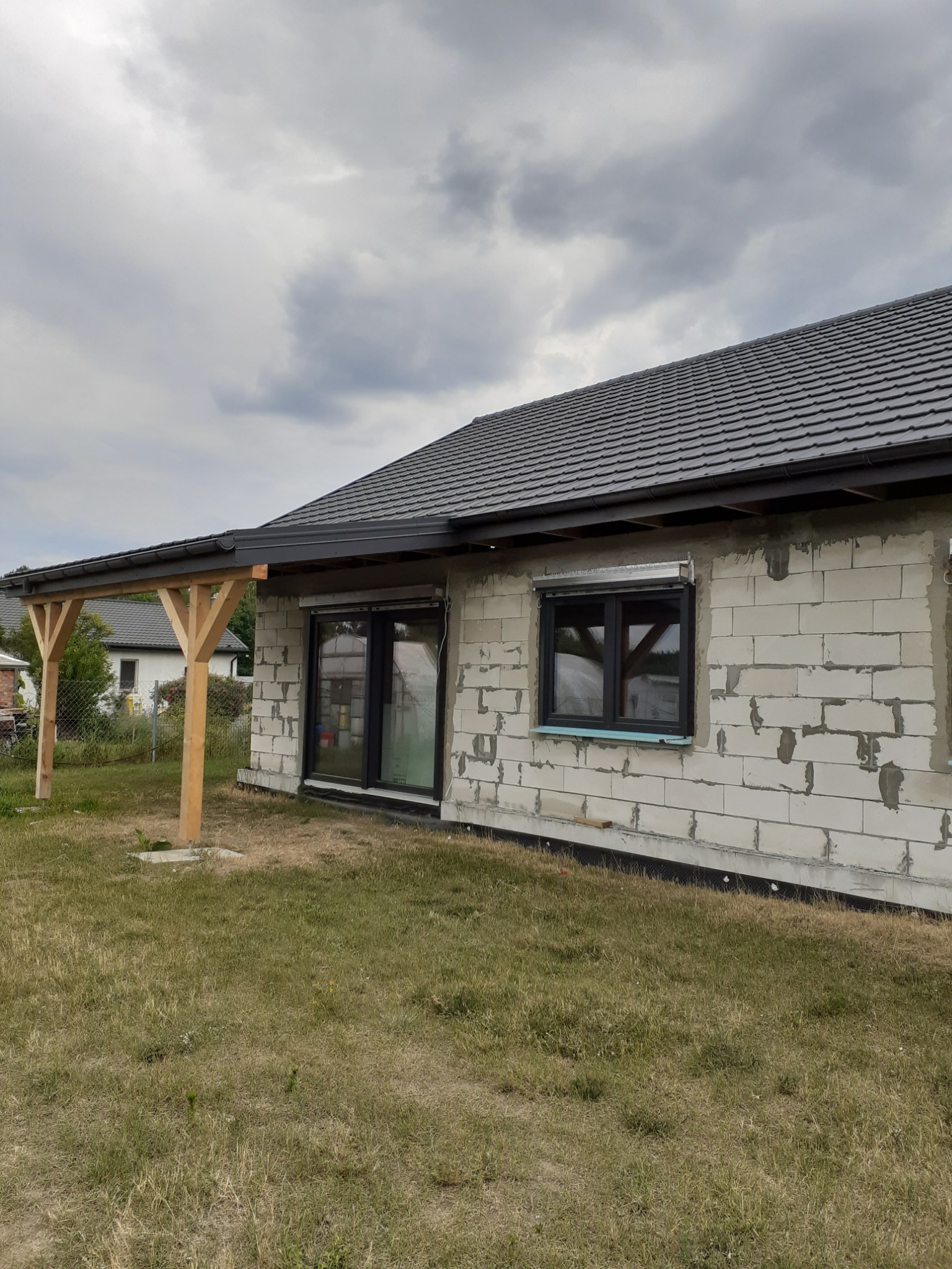 Dom w trakcie budowy z zamontowanym dachem z szarej dachówki, drewnianą konstrukcją zadaszenia tarasu i widoczną ścianą z białych bloczków.