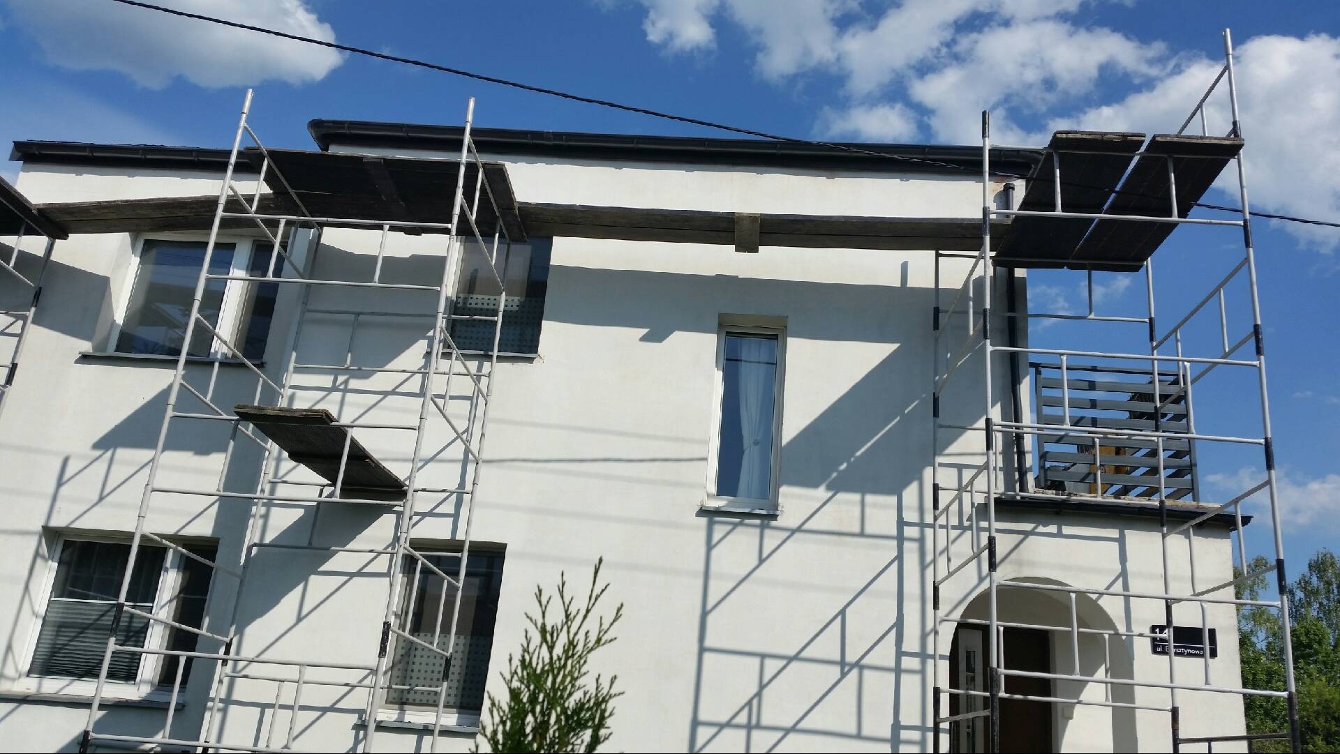 Biały budynek mieszkalny w trakcie prac remontowych, z widocznymi rusztowaniami i fragmentami dachu w trakcie wymiany, na tle błękitnego nieba z chmurami.