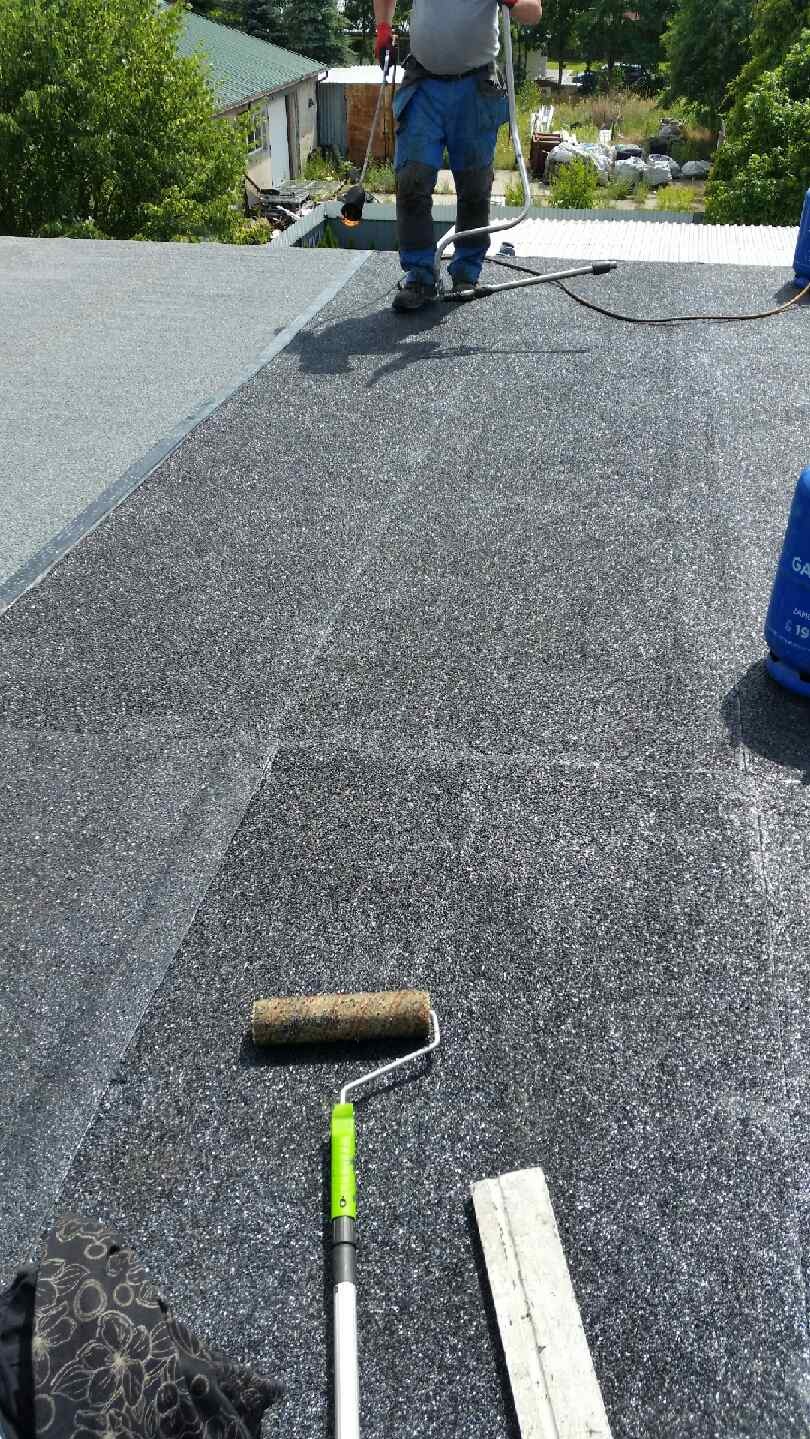 Pracownik z palnikiem dekarskim na dachu pokrytym papą, obok leży wałek do papy z zieloną rączką i deska.