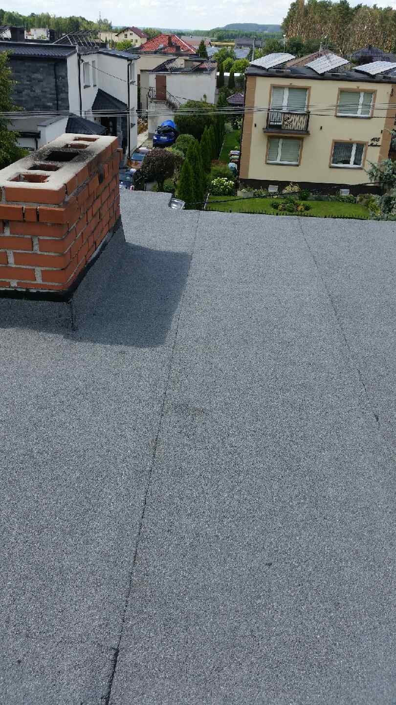 Szary dach z papy bitumicznej po kryciu, widoczny komin z cegły i zabudowania w tle.