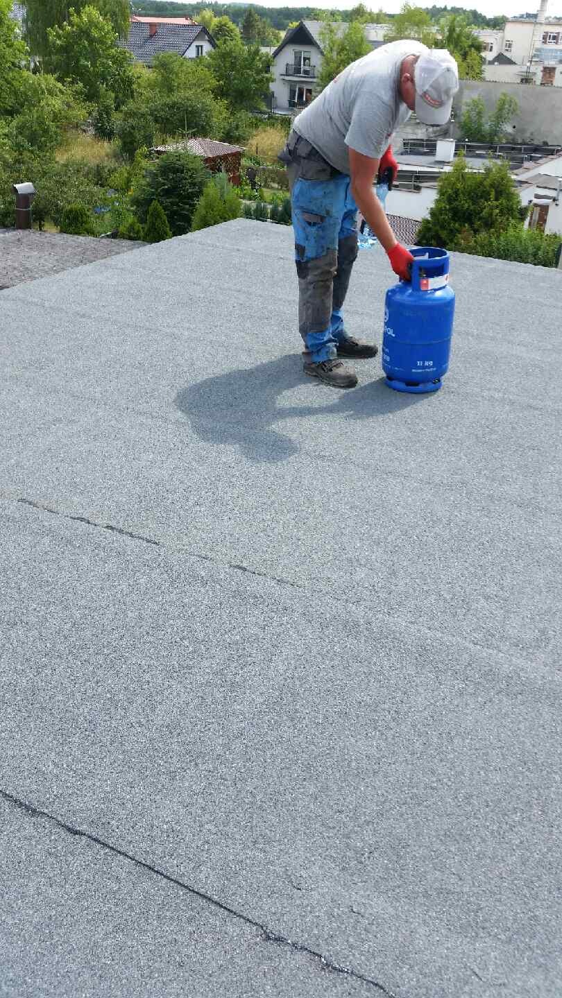 Dekarz w czerwonych rękawicach przy niebieskiej butli gazowej na pokrytym papą dachu, w tle zabudowa jednorodzinna.