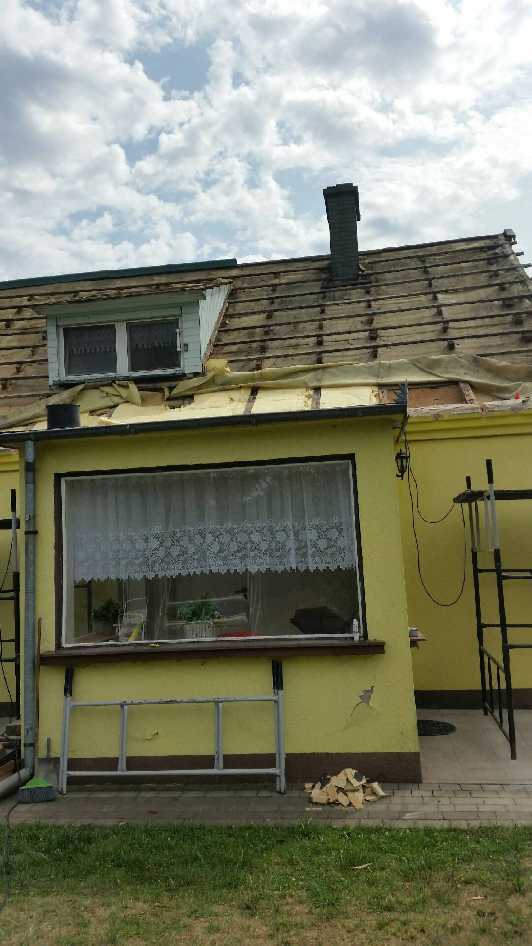 Dom w trakcie remontu dachu, widoczne fragmenty starego pokrycia, komin z cegły i okno poddaszowe.