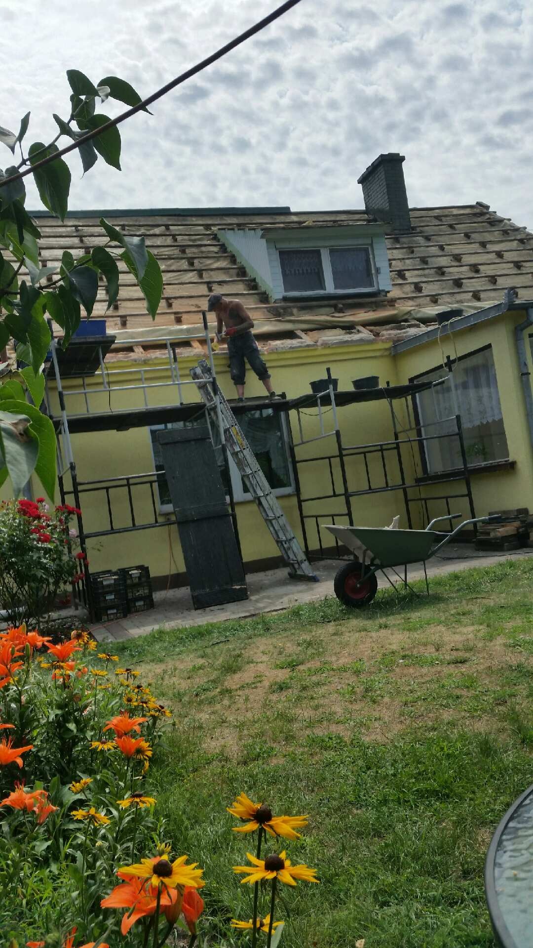 Mężczyzna bez koszulki na rusztowaniu, demontuje stare dachówki z dachu jednopiętrowego, żółtego domu z ogrodem. W tle widoczny komin i fragmenty nieba.