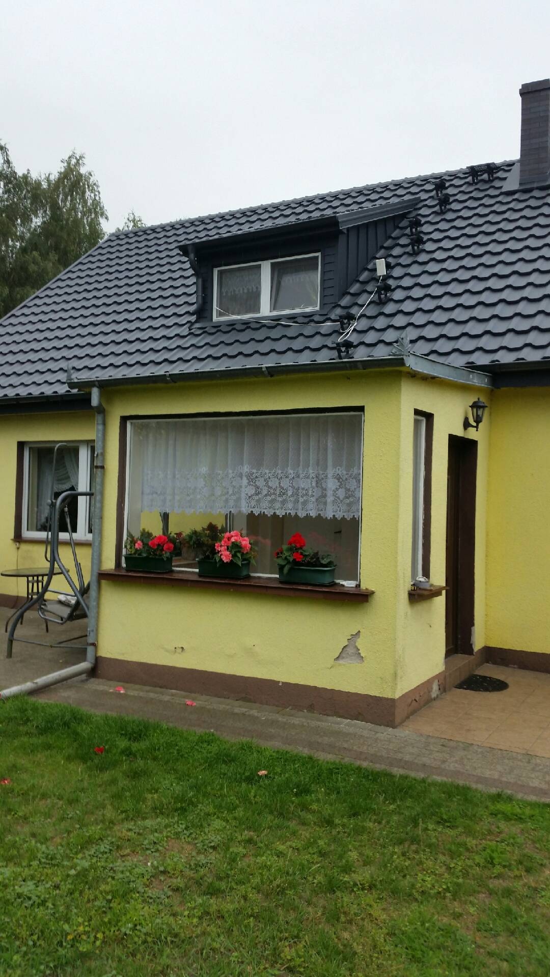 Żółty dom jednorodzinny z nowym, ciemnoszarym dachem z dachówki ceramicznej, widoczne zabezpieczenia przeciwśniegowe na połaci dachu, kwiaty w donicach na parapecie okna.