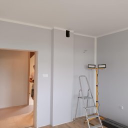 Robert Olszewski - Świeżo pomalowane na szaro wnętrze z białymi listwami przysufitowymi, widoczna drabina aluminiowa, lampa budowlana na żółtym statywie oraz fragment osoby w przejściu do innego pomieszczenia.