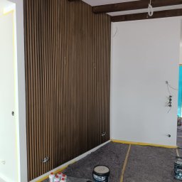 Robert Olszewski - Pokój w trakcie wykańczania z zamontowaną dekoracyjną ścianą z drewnianych paneli pionowych, widoczne puszki z farbą i akcesoria malarskie na podłodze zabezpieczonej folią ochronną oraz fragmenty...