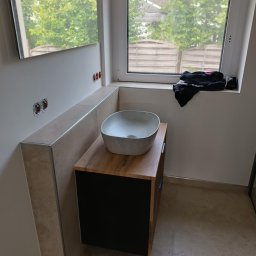 Robert Olszewski - Umywalka nablatowa w kształcie misy, zamontowana na szafce z drewnianym blatem i czarnymi frontami w łazience z beżowymi płytkami; widoczne gniazdka elektryczne i okno z widokiem na zieleń.