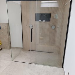 Robert Olszewski - Nowoczesna kabina prysznicowa typu walk-in z czarnymi profilami, zamontowana w łazience wykończonej płytkami imitującymi drewno i beżowym gresem.