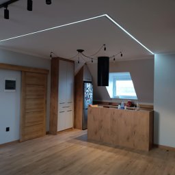 Robert Olszewski - Nowoczesne wnętrze: drewniana podłoga, wyspa kuchenna z blatem, białe szafki, przesuwne drzwi z drewnianym obramowaniem, oświetlenie LED wzdłuż sufitu i ściany, oraz designerska lampa wisząca...