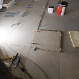 Układanie beżowych płytek podłogowych z użyciem systemu poziomowania, widoczny odpływ liniowy, wiadro z klejem i narzędzia glazurnicze.