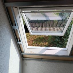 Robert Olszewski - Montaż okna dachowego w trakcie prac wykończeniowych, widoczne profile metalowe i płyty kartonowo-gipsowe, z widokiem na zielony ogród przez otwarte okno.