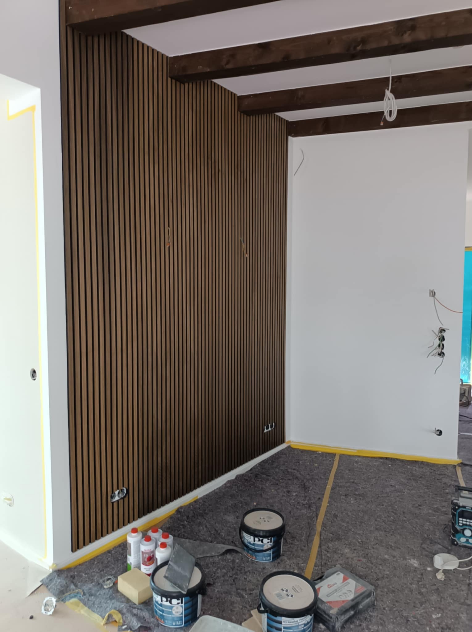 Pokój w trakcie wykańczania z zamontowaną dekoracyjną ścianą z drewnianych paneli pionowych, widoczne puszki z farbą i akcesoria malarskie na podłodze zabezpieczonej folią ochronną oraz fragmenty...