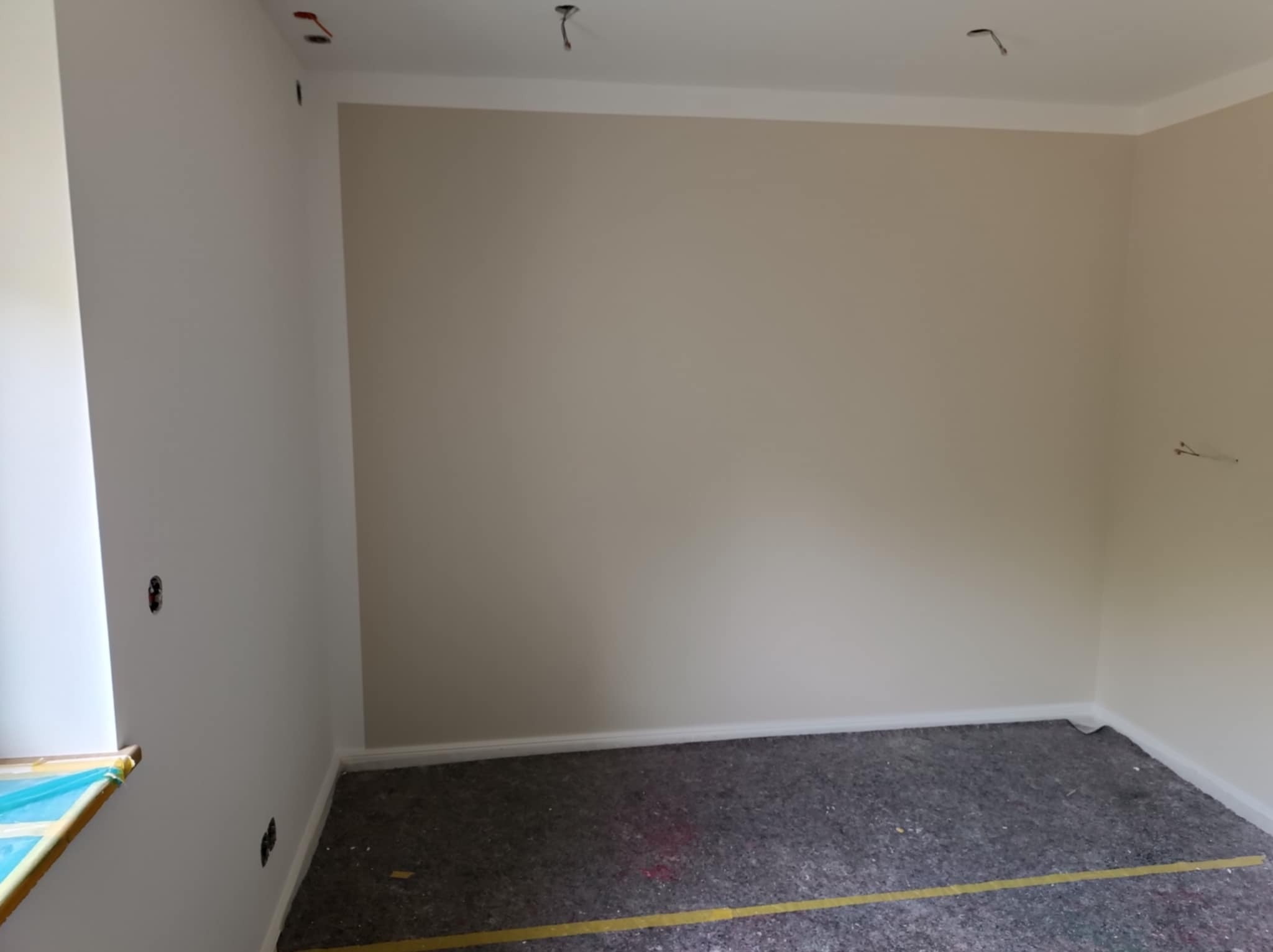 Wnętrze pokoju po świeżym pomalowaniu ścian na jasny beż, z zabezpieczoną podłogą szarą folią malarską i żółtą taśmą, widoczne wystające kable ze ściany i sufitu.