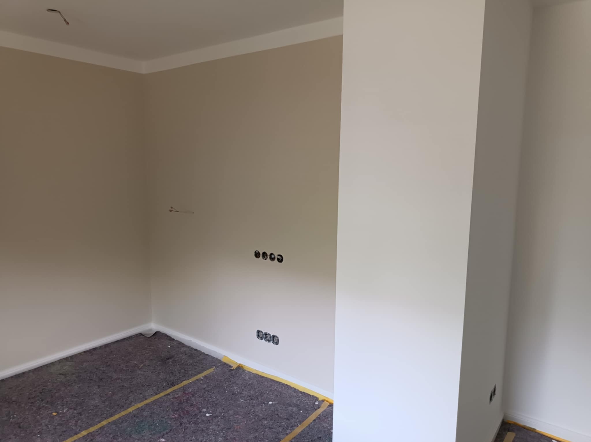 Pomieszczenie po odświeżeniu ścian w odcieniach beżu i bieli, widoczne zabezpieczenie podłogi folią malarską, gniazdka elektryczne bez osprzętu.