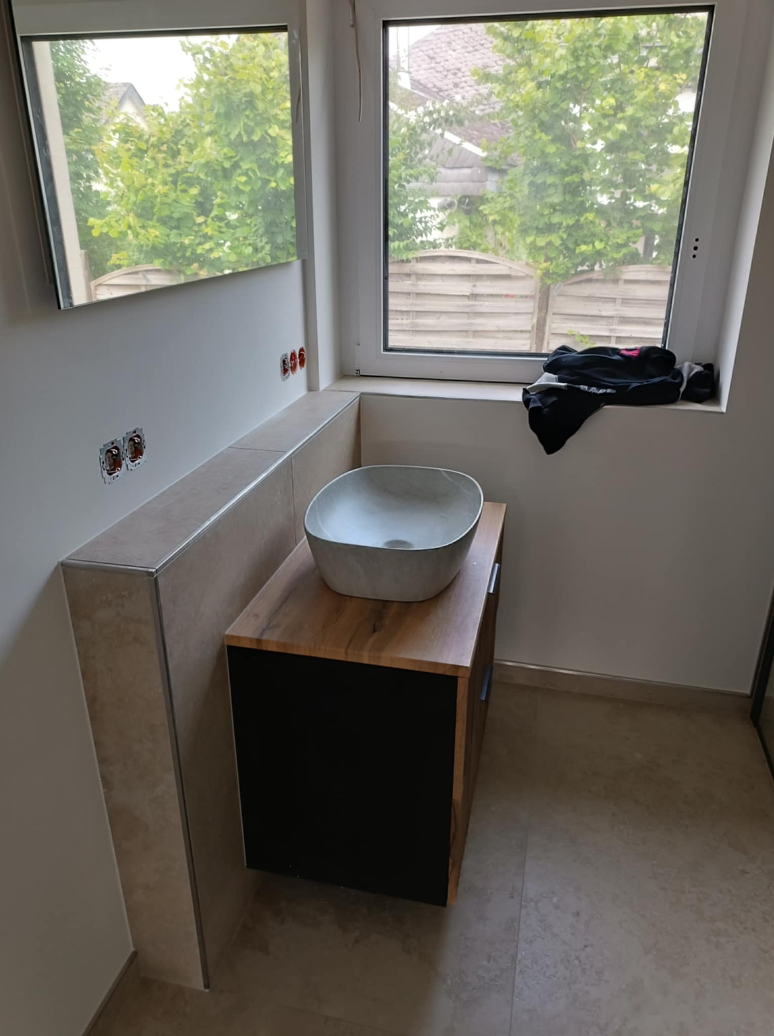 Umywalka nablatowa w kształcie misy, zamontowana na szafce z drewnianym blatem i czarnymi frontami w łazience z beżowymi płytkami; widoczne gniazdka elektryczne i okno z widokiem na zieleń.