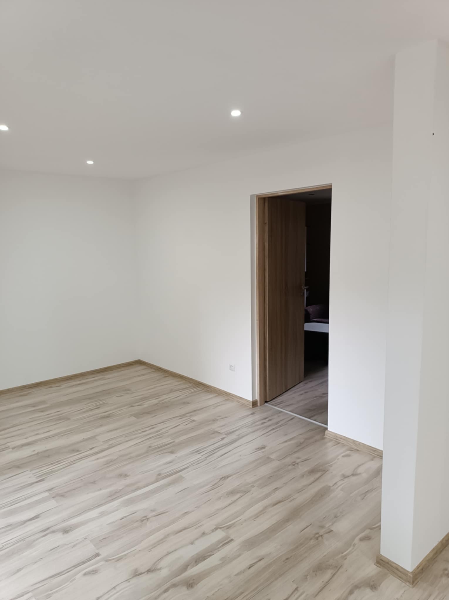 Jasne panele podłogowe ułożone w pustym, odświeżonym pokoju z białymi ścianami i otwartymi drewnianymi drzwiami w tle.