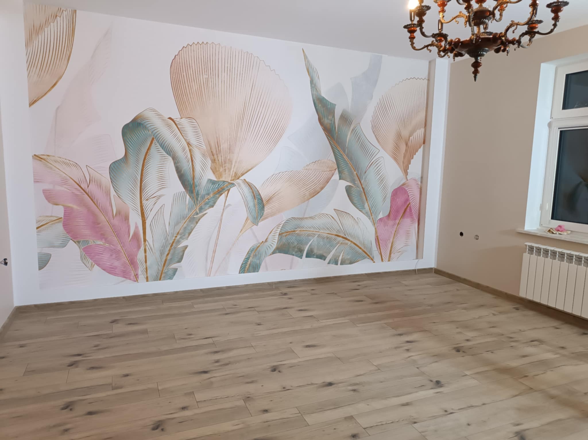 Jasne panele podłogowe w pustym pokoju z dekoracyjną tapetą w pastelowe liście na ścianie, widoczny żyrandol i grzejnik pod oknem.