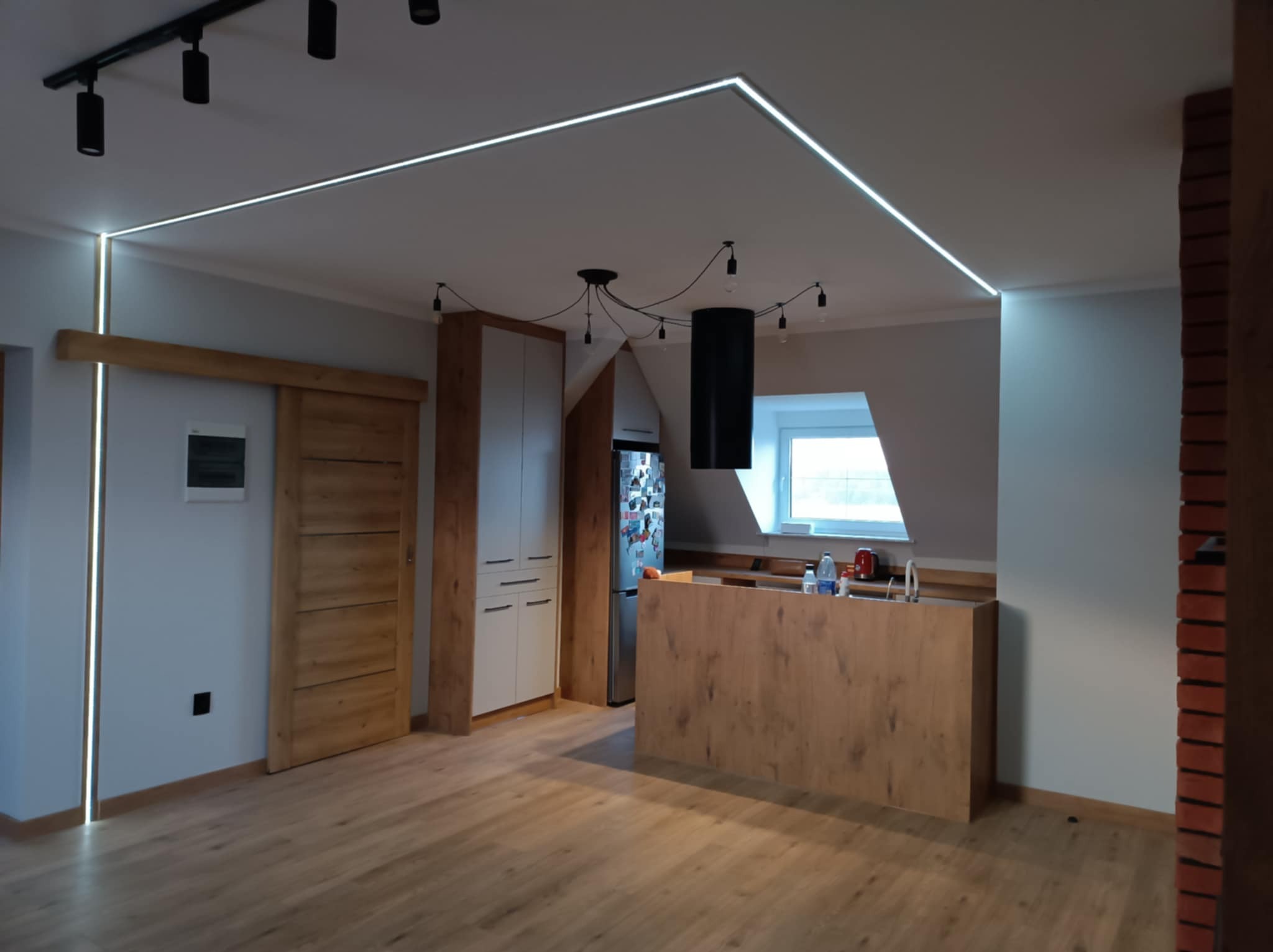 Nowoczesne wnętrze: drewniana podłoga, wyspa kuchenna z blatem, białe szafki, przesuwne drzwi z drewnianym obramowaniem, oświetlenie LED wzdłuż sufitu i ściany, oraz designerska lampa wisząca...