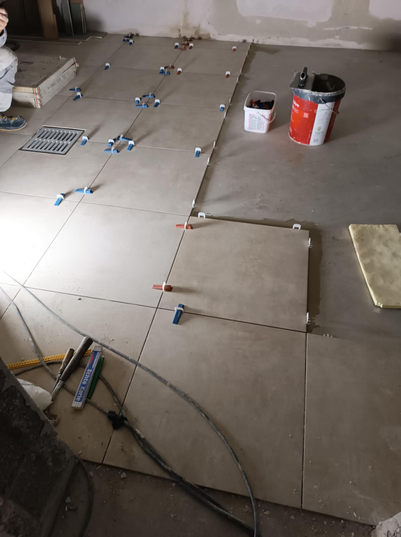 Układanie beżowych płytek podłogowych z użyciem systemu poziomowania, widoczny odpływ liniowy, wiadro z klejem i narzędzia glazurnicze.