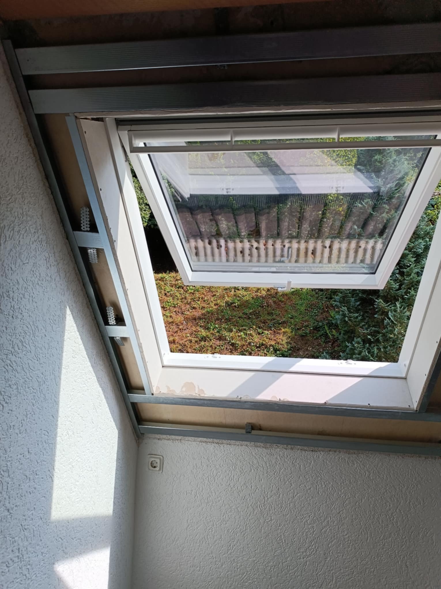 Montaż okna dachowego w trakcie prac wykończeniowych, widoczne profile metalowe i płyty kartonowo-gipsowe, z widokiem na zielony ogród przez otwarte okno.