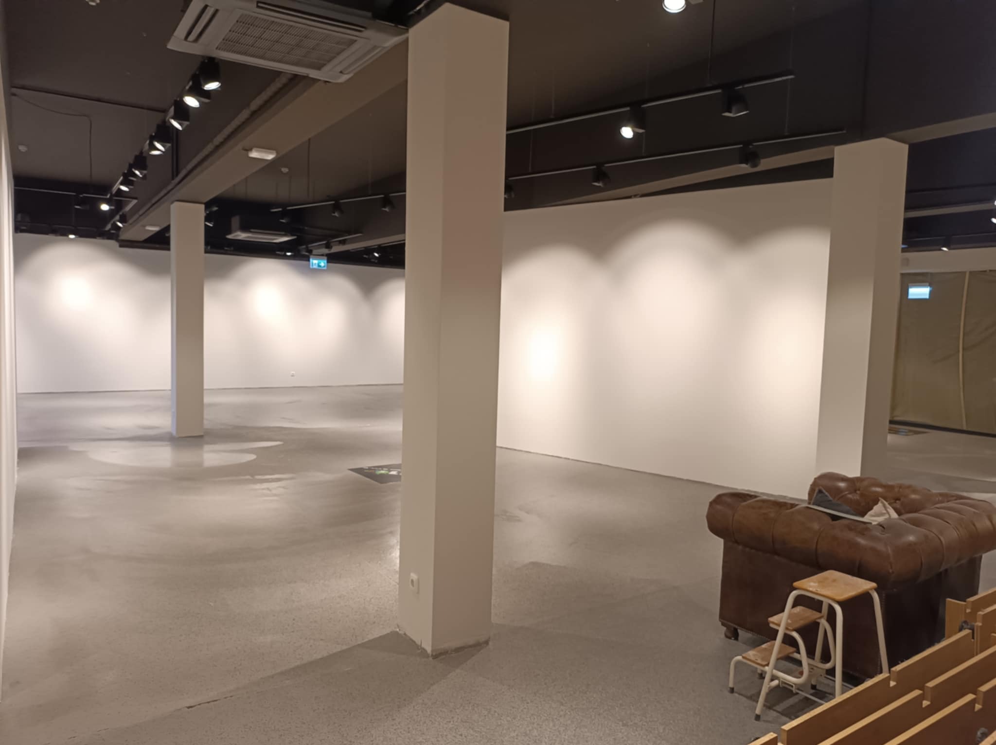 Przestronne wnętrze z surowym, betonowym podłożem, białymi ścianami i ciemnym sufitem z punktowym oświetleniem; na pierwszym planie widoczny brązowy, skórzany fotel typu chesterfield i drewniane...