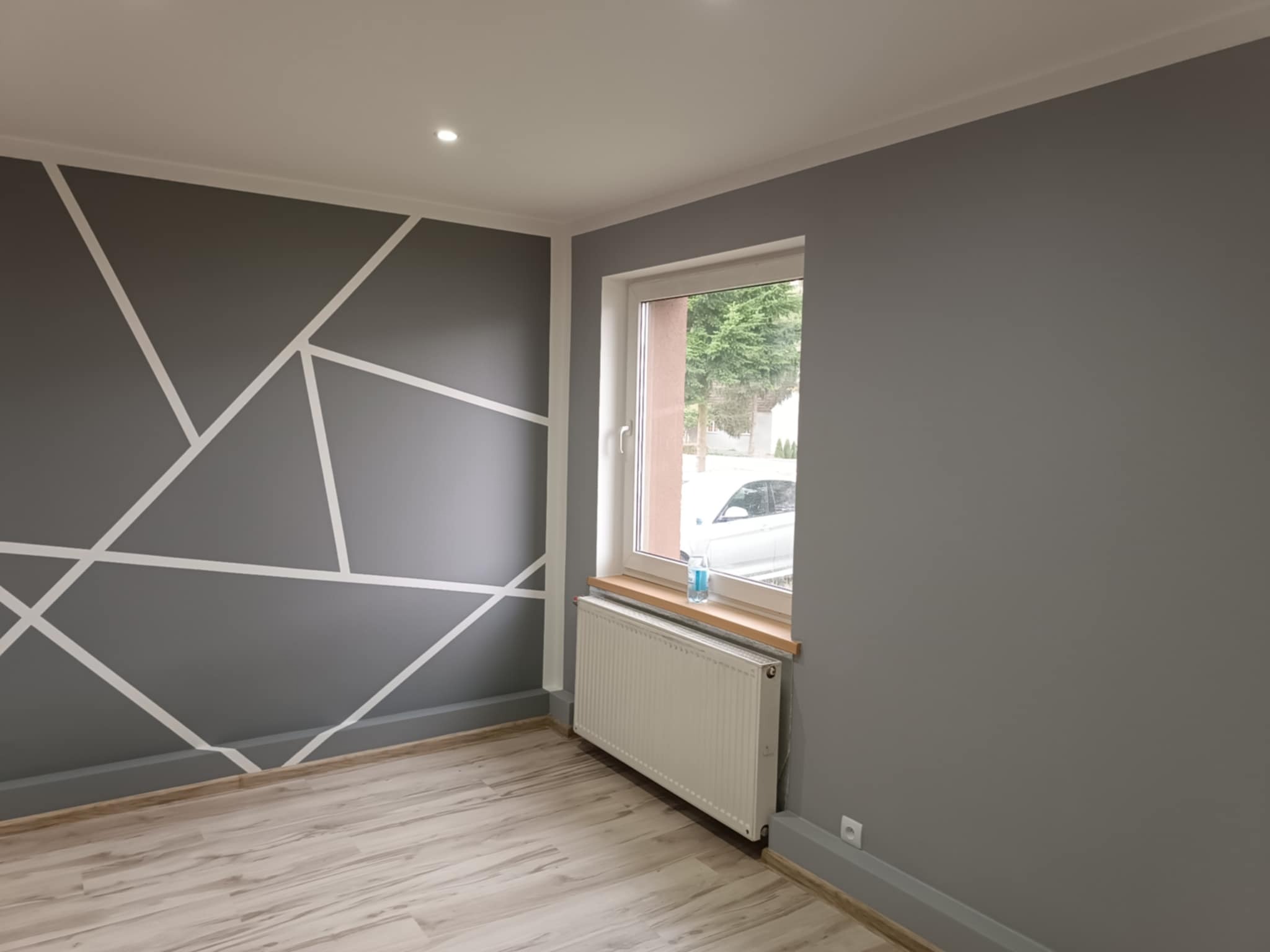 Pokój po remoncie z szarą ścianą ozdobioną białymi, geometrycznymi wzorami, drewnopodobną podłogą i białym grzejnikiem pod oknem.