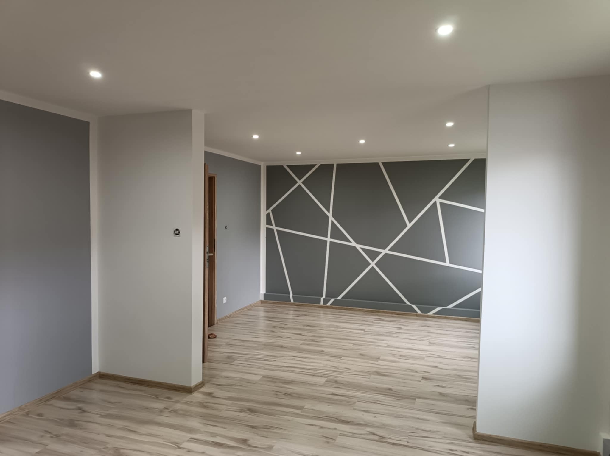 Pomalowane na szaro ściany z białymi geometrycznymi wzorami w pustym pokoju z jasnymi panelami podłogowymi i oświetleniem punktowym.