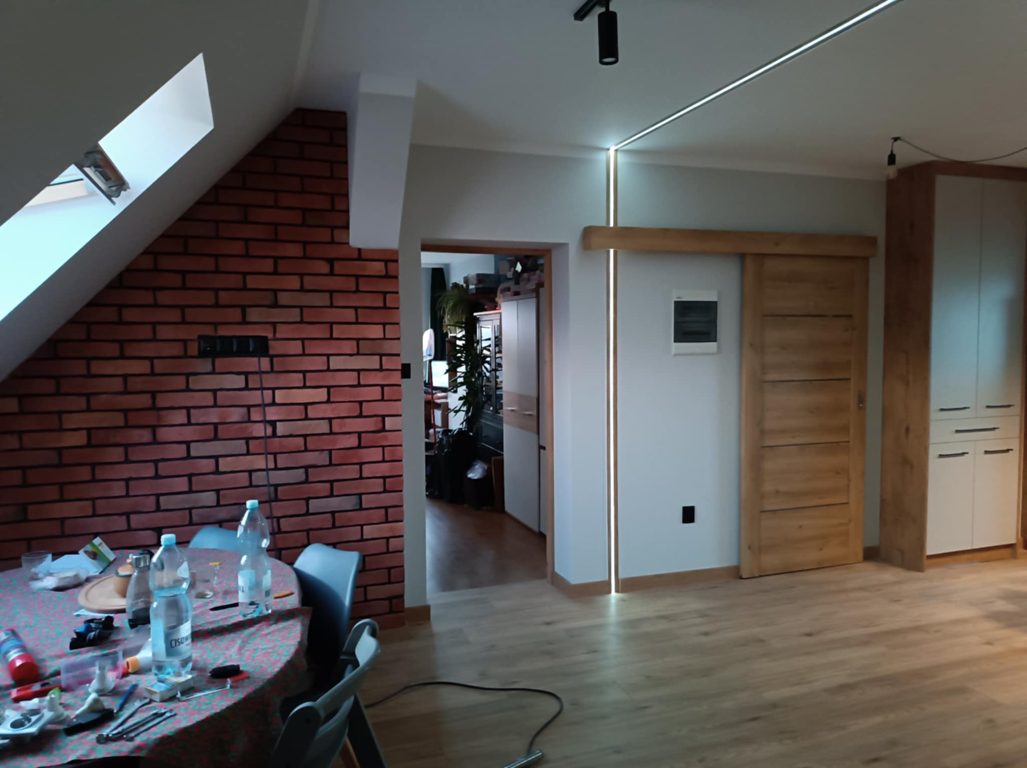 Pokój po remoncie z ceglaną ścianą, drewnianą podłogą i przesuwanymi drzwiami z drewna, widoczne elementy oświetlenia LED na suficie i przy drzwiach, stół z akcesoriami remontowymi w tle.