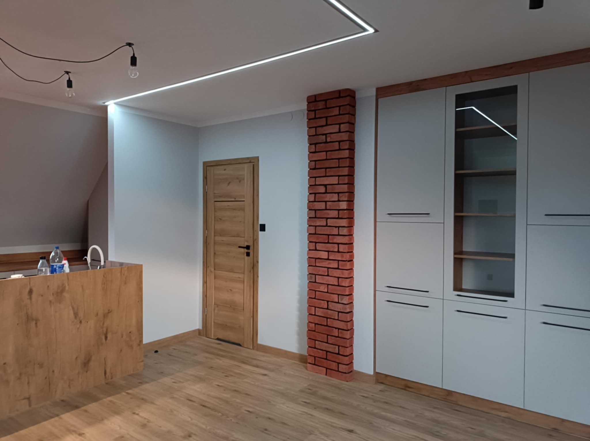 Wnętrze po remoncie w Złotowie: drewniana podłoga, białe ściany, ceglana kolumna, zabudowa meblowa z szarymi frontami i drewnianymi akcentami, drewniane drzwi i minimalistyczne oświetlenie.