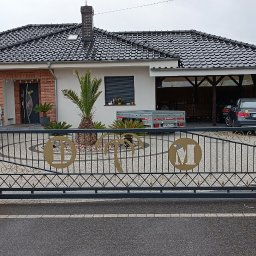 KołekSpaw - Automatyczna brama wjazdowa z ceglanymi słupkami i dekoracyjnymi elementami z motywem palm i liter, na tle domu z podjazdem i ogrodem.