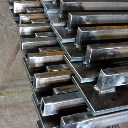 KołekSpaw - Stos metalowych elementów konstrukcyjnych, spawanych do kwadratowych podstaw, widocznych ślady spawania i obróbki metalu, w tle maszyna do cięcia metalu.