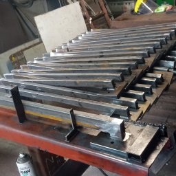 KołekSpaw - Stos metalowych profili przygotowanych do spawania, leżących na czerwonym stole warsztatowym, obok puszka ze środkiem antyodpryskowym, w tle widoczna drabina i tablica.