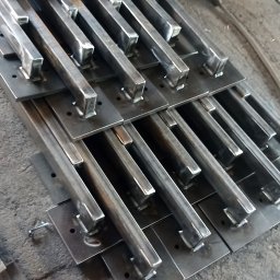 KołekSpaw - Przygotowane elementy konstrukcyjne ze stali: spawane profile kwadratowe do płaskowników z otworami, widoczny sprzęt spawalniczy w tle.