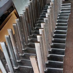 KołekSpaw - Rząd spawanych, stalowych słupków z kwadratowymi podstawami, przygotowanych do dalszej obróbki lub montażu, widoczne ślady spawania, w tle warsztatowy nieład.