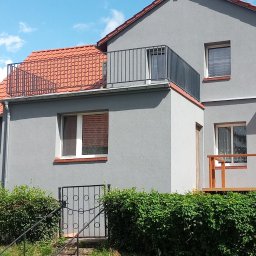 KołekSpaw - Szary dom jednorodzinny z metalową balustradą na dachu i balkonie, metalowa furtka wejściowa do piwnicy, betonowe schody, bujna zieleń, błękitne niebo.