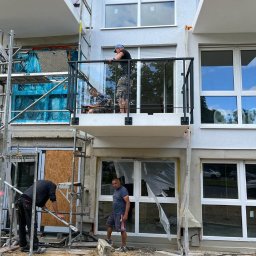 KołekSpaw - Montaż balustrady szklanej na balkonie nowego budynku, widoczni pracownicy na rusztowaniu i na balkonie, w tle okna z folią ochronną.