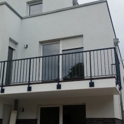 KołekSpaw - Czarna, metalowa balustrada balkonowa z pionowymi prętami, zamontowana na białym budynku z oknami z roletami zewnętrznymi, widok z dołu na pochmurne niebo.