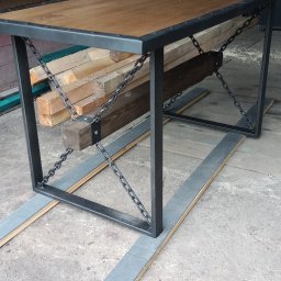 KołekSpaw - Stół z drewnianym blatem i metalową ramą wzmocnioną łańcuchami, styl industrialny, ustawiony na deskach w warsztacie.