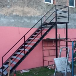 KołekSpaw - Zewnętrzne schody metalowe z platformą, prowadzące do budynku z różową ścianą, widoczne narzędzia i drabina.
