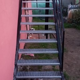 KołekSpaw - Zewnętrzne schody metalowe z kratowymi stopniami, prowadzące wzdłuż różowej ściany budynku, z widocznym tłem obejmującym budynki gospodarcze i zieleń.