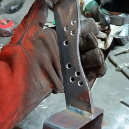 KołekSpaw - Ręcznie spawana metalowa konstrukcja z otworami, trzymana w rękawicy ochronnej na tle warsztatu.