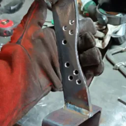 Ręcznie spawana metalowa konstrukcja z otworami, trzymana w rękawicy ochronnej na tle warsztatu.