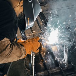 KołekSpaw - Spawanie konstrukcji metalowej: spawacz w masce ochronnej łączy elementy, iskry i dym w warsztacie.