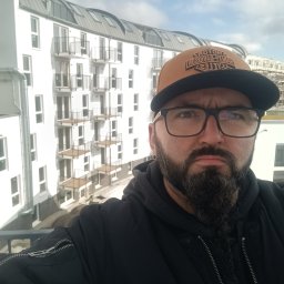 KołekSpaw - Mężczyzna w czapce i okularach robi selfie na tle nowego budynku z balkonami i balustradami.