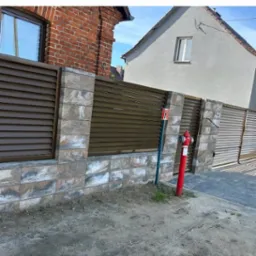 Nowoczesne ogrodzenie posesji z brązowymi, poziomymi panelami i kamienną podmurówką. Czerwony hydrant na pierwszym planie, budynki w tle.